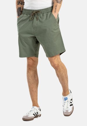 Pantalones cortos de color verde oliva con cintura elástica y cordón. Presenta bolsillos laterales y un corte relajado. Se lleva con zapatillas blancas y negras.