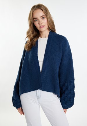 Tummansininen neulecardigan, jossa on leveä avorintus, tekstuurilla varustetut hihat, joissa on letitetty kuvio, ja ribbireunus kauluksessa. Päällä vaalea toppi.