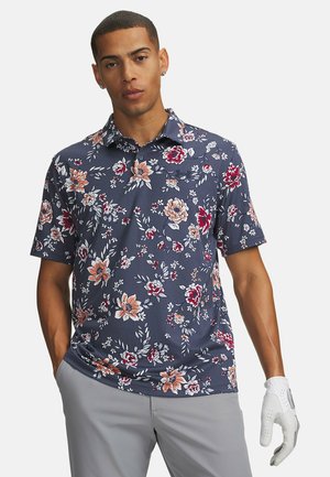 Mann trägt ein marineblaues Poloshirt mit Blumenmuster, hellgraue Hose und einen weißen Golfhandschuh, eine Hand in der Tasche.