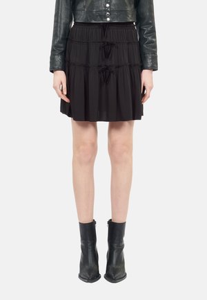 Frau trägt einen schwarzen Stufen-Minirock mit Kordelzügen, eine schwarze, kurze Lederjacke und schwarze Stiefeletten, steht vor weißem Hintergrund.