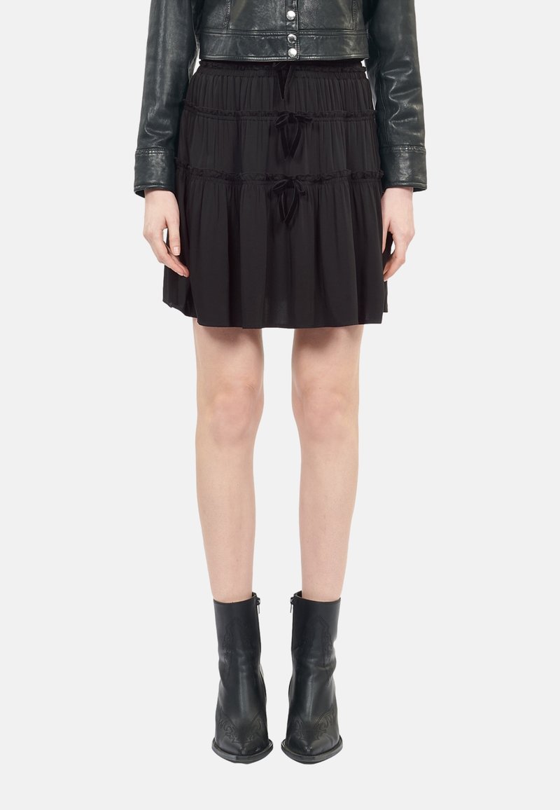 Femme portant une mini-jupe noire à volants avec des nœuds coulissants, une veste en cuir noire courte et des bottines noires, debout devant un fond blanc.
