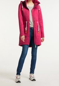 Fleece-lined pink coat with zip closure, drawstring accents, and large front pockets, paired with blue jeans and sporty shoes.