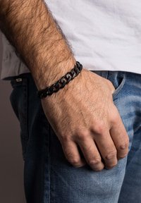 Juodas metalinis grandinės apyrankė su tarpusavyje susijungiančiomis jungtimis, dėvima ant rankos. Ranką atsiremiama į džinsinį audinį, akcentuojant tekstūrą ir spalvų kontrastą.
