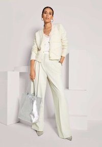 Frau in cremfarbener Strickjacke, weißem Spitzentop, weitem Hosenanzug, silbernen spitzen Schuhen und silberner Tragetasche steht selbstbewusst vor einem weißen Hintergrund.