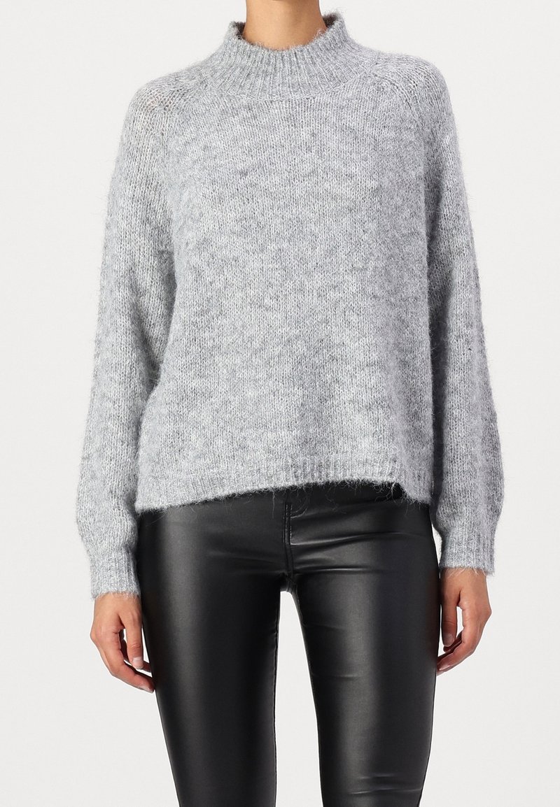 Femme portant un pull en maille pelucheux gris clair à manches longues, associé à un pantalon en cuir noir ajusté, visible des épaules aux hanches.