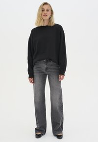 Černá oversized mikina s kulatým výstřihem, spárovaná s vysokým pasem, volnými šedými džíny s vybledlým efektem. Modelka nosí černé ploché boty.