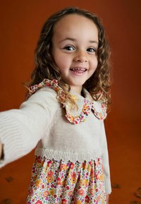 Lächelndes junges Mädchen mit lockigem Haar trägt ein Blumenkleid und einen beigen Pullover mit Rüschenkragen und streckt sich gegen einen orangefarbenen Hintergrund nach vorne.