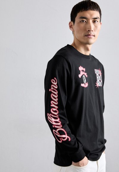 Young man wearing a black long-sleeve shirt with pink "Billionaire" text on the sleeve and graphic designs on the chest, hands in pockets.