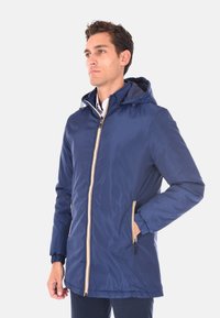 Uomo che indossa una giacca a cappuccio blu navy con una cerniera a righe bianche e gialle, in piedi con una mano in tasca, mentre guarda di lato.