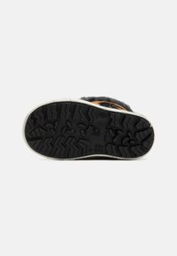 Schwarze Gummisohle mit sichtbarem Profilmuster und beigen Kanten. Weiches schwarzes Obermaterial mit subtilen Branding-Details nahe der Ferse.