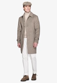 Brun houndstooth-frakke med snitkrave, beige rullekrave, hvide bukser og brune snørebåndsstøvler. Kasketten tilføjer et klassisk præg.