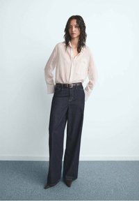 Vrouw met golvend donker haar, gekleed in een beige gestreepte blouse met knopen, in een hoge taille gestoken in donkere, wijde jeans met hoge taille en bruine puntige schoenen.