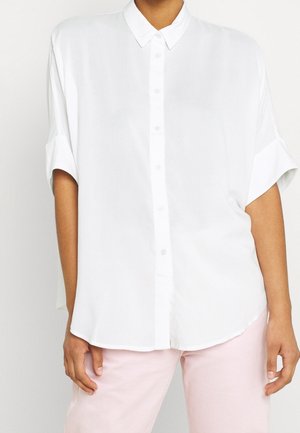 Personne portant une chemise blanche ample à manches courtes boutonnée et un pantalon rose clair sur un fond clair uni.