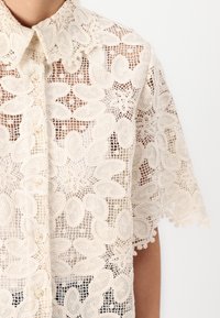 Blusa in pizzo avorio con motivi floreali, maniche corte, colletto e chiusura a bottoni. Presenta ricami intricati e una tessitura aperta.