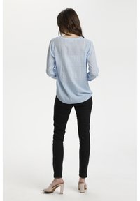 Blouse à manches longues bleu clair avec un motif subtil en losanges, coupe décontractée, associée à un jean skinny noir et des sandales à talons blocs couleur nude.