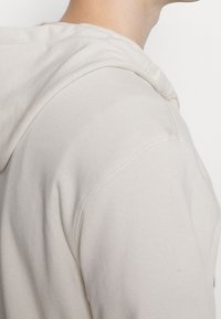 Felpa allacciata di un beige chiaro, realizzata in tessuto morbido con una vestibilità comoda. Presenta un cappuccio con coulisse e cuciture a vista lungo le giunture.