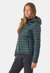 Donkergroene pufferjack met horizontale stiknaad, capuchon en voorzakken met rits. Draagover een zwarte top, gecombineerd met donkere broek.