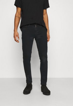 Man met zwarte slim-fit jeans, zwarte suède laarzen en een zwart T-shirt met korte mouwen, staand tegen een effen lichtgrijze achtergrond.