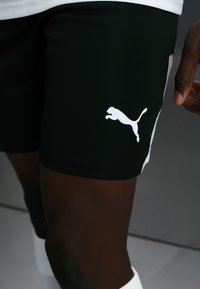 Άτομο που φοράει μαύρα σορτς Puma με λευκό λογότυπο Puma στον μηρό, στέκεται σε ουδέτερο φόντο.