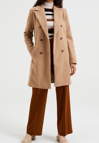Beige double-breasted coat with notched lapels, featuring rounded buttons and a straight silhouette, paired with brown wide-leg trousers.