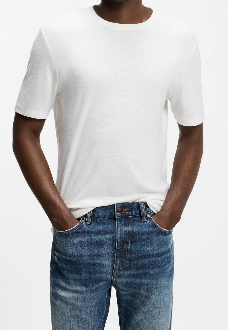 Homme portant un t-shirt blanc uni à manches courtes et un jean en denim bleu, les mains dans les poches, sur fond blanc.