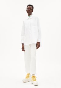 White long-sleeve shirt with button closure and pocket, paired with white cropped pants and chunky white sneakers with yellow laces.