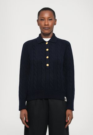 Maglione polo lavorato a maglia blu navy con dettagli a treccia, dotato di orlo a coste, cinque bottoni dorati e maniche lunghe. Indossato sopra pantaloni neri.