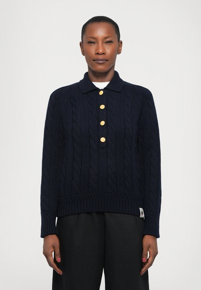 Polo en tricot navy avec des détails en cable, doté d'un ourlet à côtes, de cinq boutons dorés et de manches longues. Porté sur un pantalon noir.