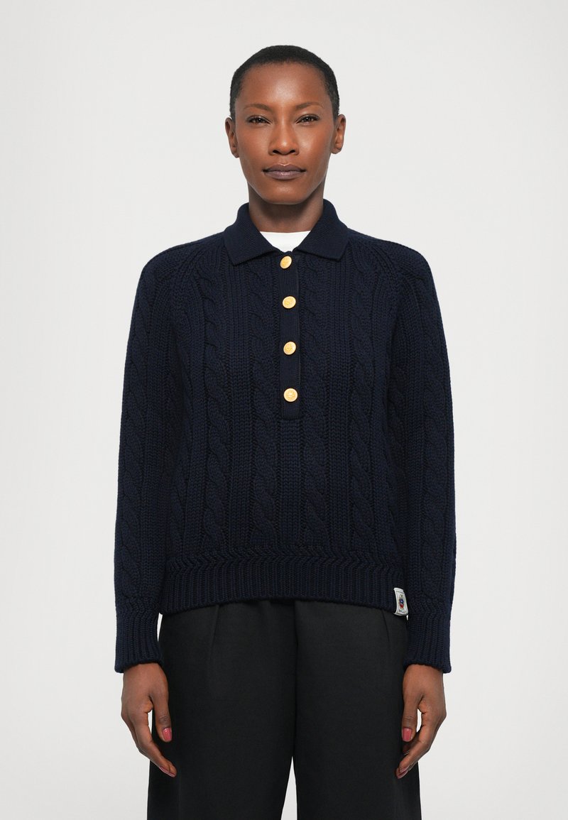 Polo en tricot navy avec des détails en cable, doté d'un ourlet à côtes, de cinq boutons dorés et de manches longues. Porté sur un pantalon noir.