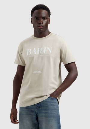 Jonge man staand met één hand in zijn zak, draagt een beige T-shirt met witte tekst en een blauwe spijkerbroek, neutrale grijze achtergrond.