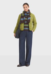 Veste matelassée verte sur un pull marron rayé, associée à un pantalon en denim noir à jambes larges et une écharpe assortie. Sac bandoulière noir.
