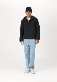 Chaqueta negra con capucha, cierre frontal y bolsillos, llevada sobre una camisa blanca, combinada con jeans de mezclilla azul claros y zapatillas blancas. Gorra negra con logo.