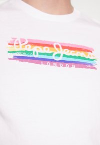 Camiseta blanca de algodón con la inscripción "Pepe Jeans" en cursiva y rayas de colores en tonos del arcoíris, y "LONDON" debajo en una fuente más sencilla.