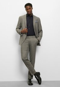 Grauer Karoanzug mit schmaler Passform, kombiniert mit einem dunklen Hemd. Schwarze Lederschuhe runden das Outfit ab und zeigen ein dezentes Einstecktuch-Detail.