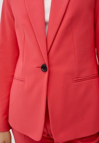 Blazer en corail avec une texture lisse, fermeture à un bouton, et deux poches avant, présentant une coupe ajustée et des revers aigus.