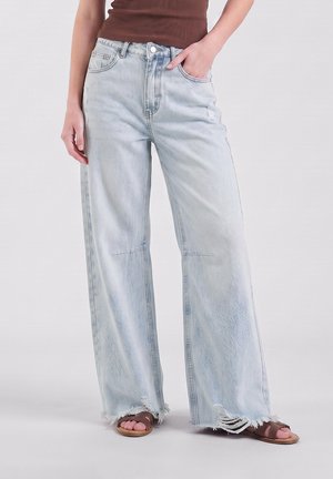 Persona che indossa jeans azzurri a gamba larga con orli sfrangiati e sandali marroni, mano in tasca, su sfondo semplice.