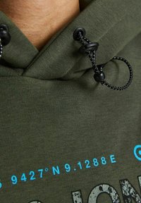 Sudadera verde oliva hecha de un tejido suave, que cuenta con un cordón con acentos negros y texto azul que muestra coordenadas y gráficos.