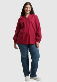 Rote, strukturierte Bluse mit langen Ärmeln, V-Ausschnitt und Knopfleiste, kombiniert mit blauen Jeans und weißen Turnschuhen.