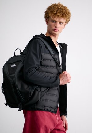 Joven con pelo rizado que lleva una chaqueta negra y pantalones rojos, con una mochila negra North Face en un hombro contra un fondo blanco.