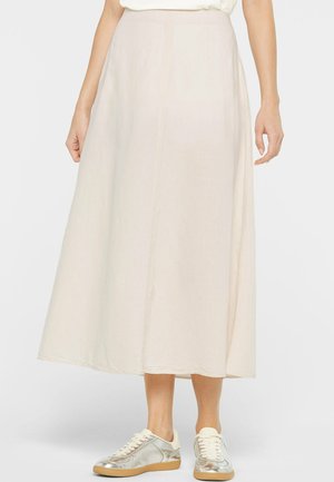 Frau trägt einen cremefarbenen Midi-Rock kombiniert mit metallisch silbernen Sneakers, steht vor einem einfarbig hellen Hintergrund.