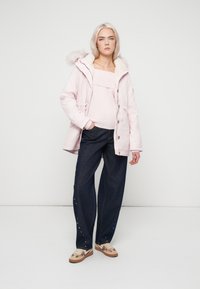 Mulher com cabelo loiro a usar uma jaqueta com capuz rosa claro, suéter rosa pálido, calças largas escuras com pequenos adornos e sapatilhas bege.