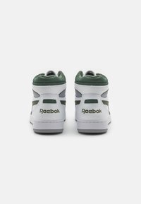 Vita hög-top Reebok sneakers med gröna detaljer. Funktioner inkluderar en texturerad meshkrage, grå ränder och logotyp på hälen.