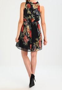 Blommönstrad, ärmlös svart klänning med plisserad kjol, rund hals och elastisk midja. Buren med svarta högklackade skor.