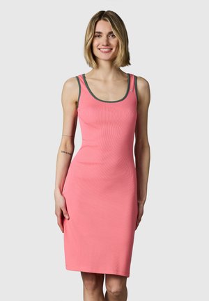 Lachende vrouw met blond haar, die een mouwloze roze jurk met donkere bies draagt, staand voor een lichtgrijze achtergrond.