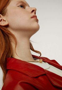 Collar de color dorado con un colgante esférico texturizado, usado sobre una prenda blanca y una chaqueta de cuero roja. El fondo es neutro.