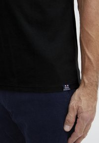 Fekete pamut póló egyenes lefelé álló szegéllyel, az alján bal oldalon egy kis kék és fehér címkével. Az anyaga simának tűnik.