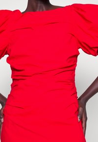 Frau in einem leuchtend roten Kleid mit Puffärmeln und Raffung, mit auf Hüften platzierten Händen vor einem schlichten Hintergrund.