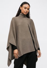 Γυναίκα φορώντας μια καφέ τζάκετ με ζιβάγκο πάνω από ένα μαύρο μπλουζάκι μακροχρόνιου μανικιού και μαύρο παντελόνι, στέκεται μπροστά σε ένα απλό λευκό φόντο.