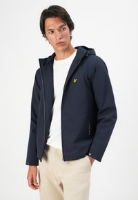 Man wearing navy blue hooded jacket over white shirt and beige pants, looking sideways with one hand in pocket against white background.
