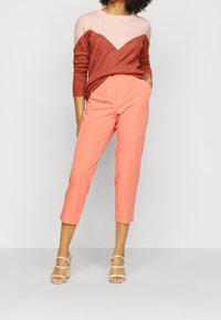 Sweter w blokowe wzory w kolorze czerwonym i różowym z wzorem w kształcie litery V, połączony z koralowymi krótkimi spodniami i beżowymi sandałami na obcasie z paskami. Gładkie tekstury na całej powierzchni.